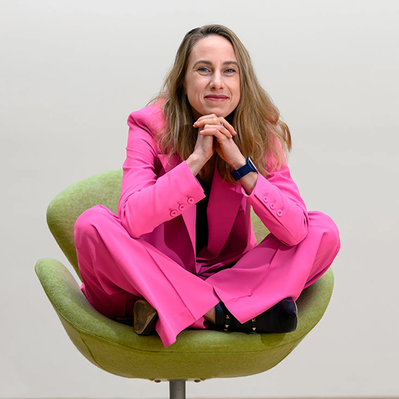 Polina Anikeeva in a pink suit, sitting cross-legged in a chair.