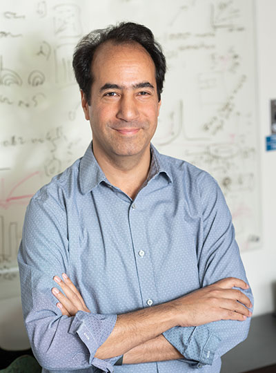 A man stands with his arms crossed in front of a board with mathematical equations written on it.