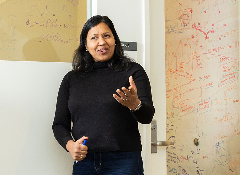 Woman gestures with her hand in front of a glass wall with equations written on it.