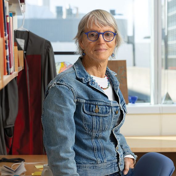 Female researcher leaning against desk with window in background.